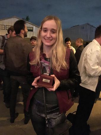 Me (Chiara Gatti), a young white woman with blonde hair holding a trophy shaped like a dolphin. The dolphin is made of purple felt and the base it is on is wood with a metal plaque.