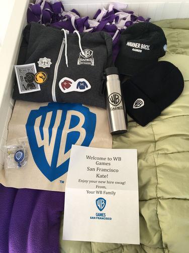 A photo of new employee swag given to new WB Games San Francisco employees. There is a hoodie, a baseball cap, a beanie, a water bottle, a tote bag, several stickers, a lanyard/badge holder clip, and a note that reads "Welcome to WB Games San Francisco Chiara! Enjoy your new hire swag! From, Your WB Family". Under the words is the logo for WB Games San Francisco in blue. The logo is a shield with the letters W and B inside it, and beneath the shield is the word "GAMES". Under that is "SAN FRANCISCO".